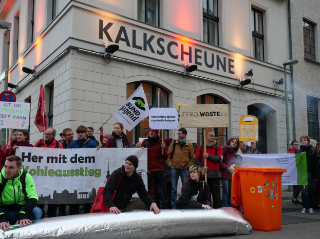 Eine Gruppe von Menschen demonstriert vor einem Gebäude in Deutschland, mit Schildern und Plakaten, während zwei Personen im Vordergrund sitzen und ein Müllcontainer rechts zu sehen ist.