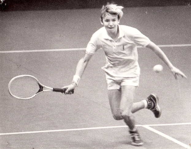 Ein junger Mann in weißer Tennisbekleidung hält einen Schläger und bereitet sich auf den Schlag eines Tennisballs auf einem Platz vor.