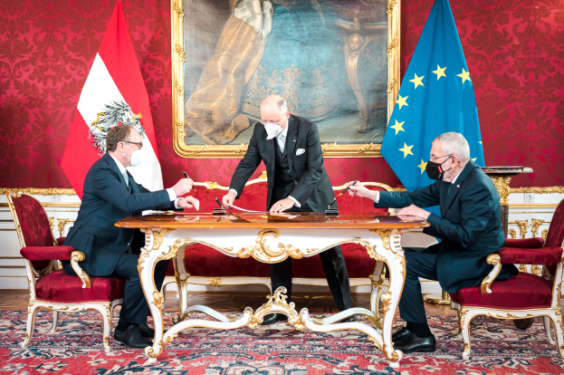 Drei Männer in Mänteln und Masken sitzen an einem Tisch mit Papieren und Gegenständen vor zwei Flaggen, mit einem Foto Rahmen an der Wand dahinter, der wie ein Treffen zwischen dem österreichischen Bundeskanzler und dem Präsidenten der Europäischen Union aussieht.