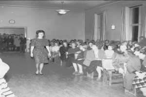 Eine Frau in einem schwarzen und weißen Kleid läuft über den Laufsteg während einer Modenschau für Frauenrechte, mit einer Menge, die auf Stühlen sitzt und einer Uhr und Lichtern an der umliegenden Wand.
