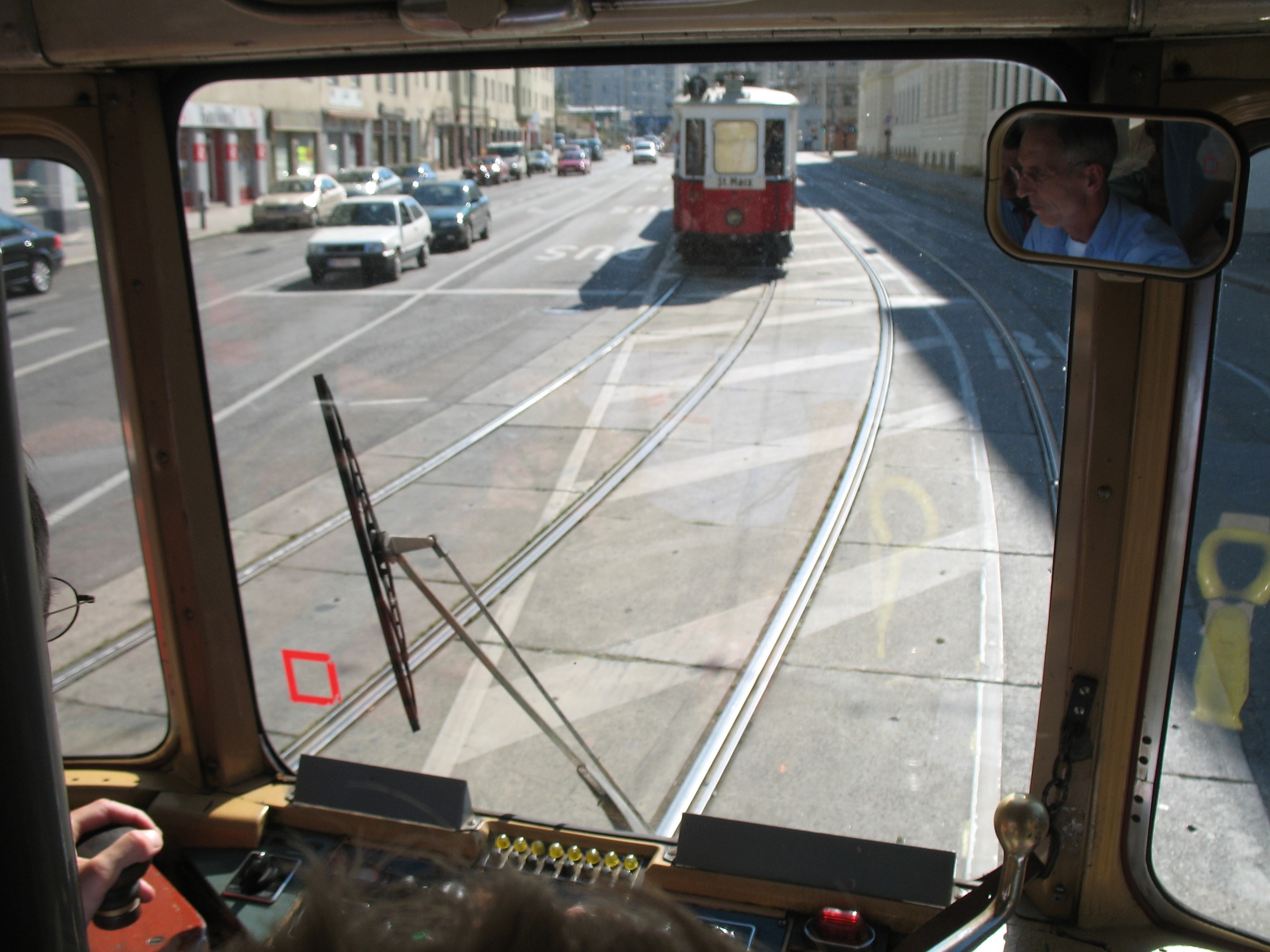 Ein Mann fährt eine Tram auf einer Stadtstraße mit anderen Fahrzeugen, Gebäuden und einem klaren blauen Himmel im Hintergrund.