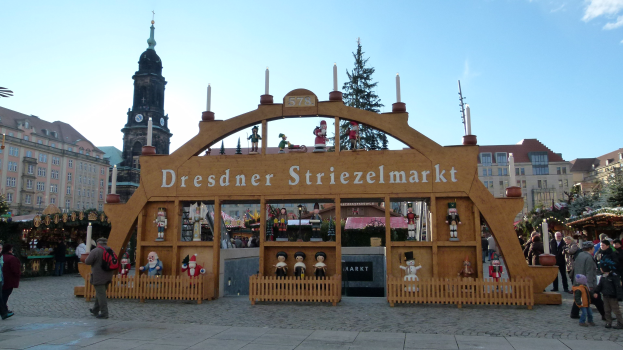 Ein belebter Weihnachtsmarkt in Dresden, Deutschland, mit Menschen, die herumlaufen, einige mit Taschen, unter einem Bogen mit Text, Bäumen und Gebäuden mit Fenstern im Hintergrund unter einem bewölkten Himmel.