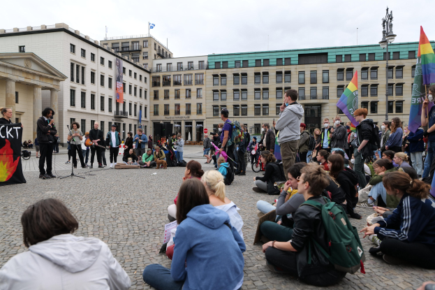 Eine Gruppe von Menschen, die auf dem Boden vor einer Menge sitzt, die Fahnen und Transparente hält, mit einer Person, die in ein Mikrofon spricht, einer Statue und Gebäuden im Hintergrund während einer Homo-Protest in Berlin, Deutschland.