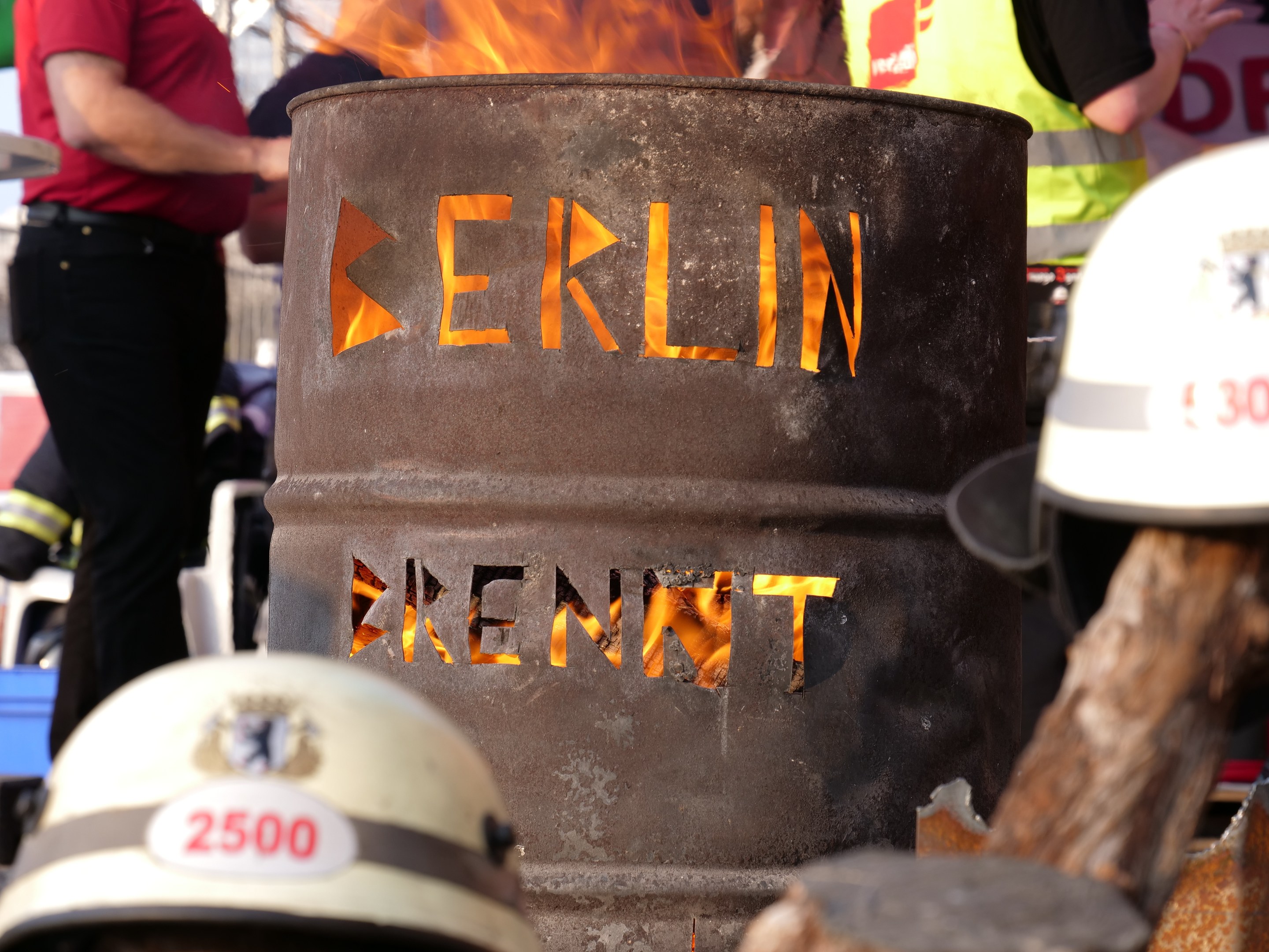 Eine Gruppe von Menschen, die um einen Feuerhydranten mit der Aufschrift 'Berlin' stehen, mit einem brennenden Fass im Vordergrund, Helme unten und einem hölzernen Objekt auf der rechten Seite.