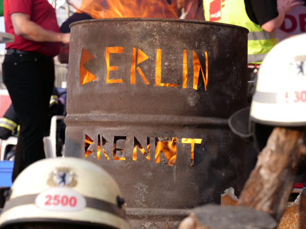 Eine Gruppe von Menschen, die um einen Feuerhydranten mit der Aufschrift 'Berlin' stehen, mit einem brennenden Fass im Vordergrund, Helme unten und einem hölzernen Objekt auf der rechten Seite.
