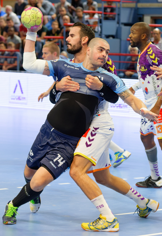 Eine Gruppe von Männern, die in einem Stadion Handball spielen, wobei einer einen Ball hält und Zuschauer im Hintergrund sitzen.