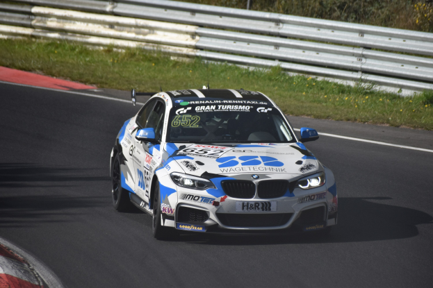 Ein BMW M4 DTM-Rennwagen mit Text und Zahlen auf der Karosserie, der auf einer von Gras umgebenen Rennstrecke mit einem Zaun und Pflanzen im Hintergrund fährt.