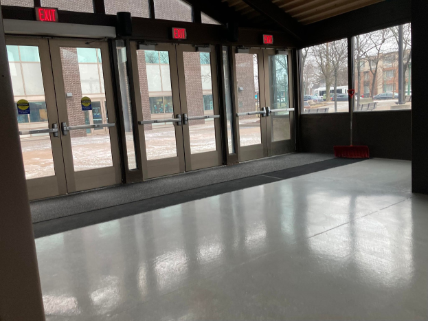 Ein großer Raum mit vielen Fenstern und Türen, ein Besen auf dem Boden, Schilder, ein Dach, durch Fenster sichtbare Bäume, Fahrzeuge und Gebäude draußen und ein bewölkter Himmel, was auf einen kommerziellen Bodenreinigungsdienst hinweist.