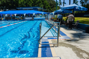 Ein großes Schwimmbad mit schwimmenden Menschen, umgeben von Geländern, Sonnenschirmen, Stühlen, Tischen, Pflanzen, Gras, Bäumen und einem Gebäude unter einem klaren blauen Himmel.