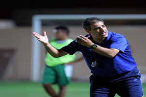 Ein Mann in einem blauen T-Shirt und blauen Hosen steht auf einem Fußballfeld und hält ein Mikrofon in der Hand, während er zu seinen Spielern gestikuliert. Im Hintergrund ist eine Person in einem grünen Kleid, ein Torpfosten und eine Wand zu sehen.