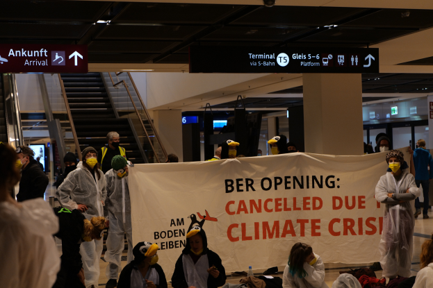 Eine Gruppe maskierter Individuals mit einer Tafel, auf der "Ber Opening Due to Climate Crisis" steht, umgeben von verstreuten Taschen und Gegenständen und im Hintergrund Rolltreppen, Texttafeln, Lampen und Säulen.