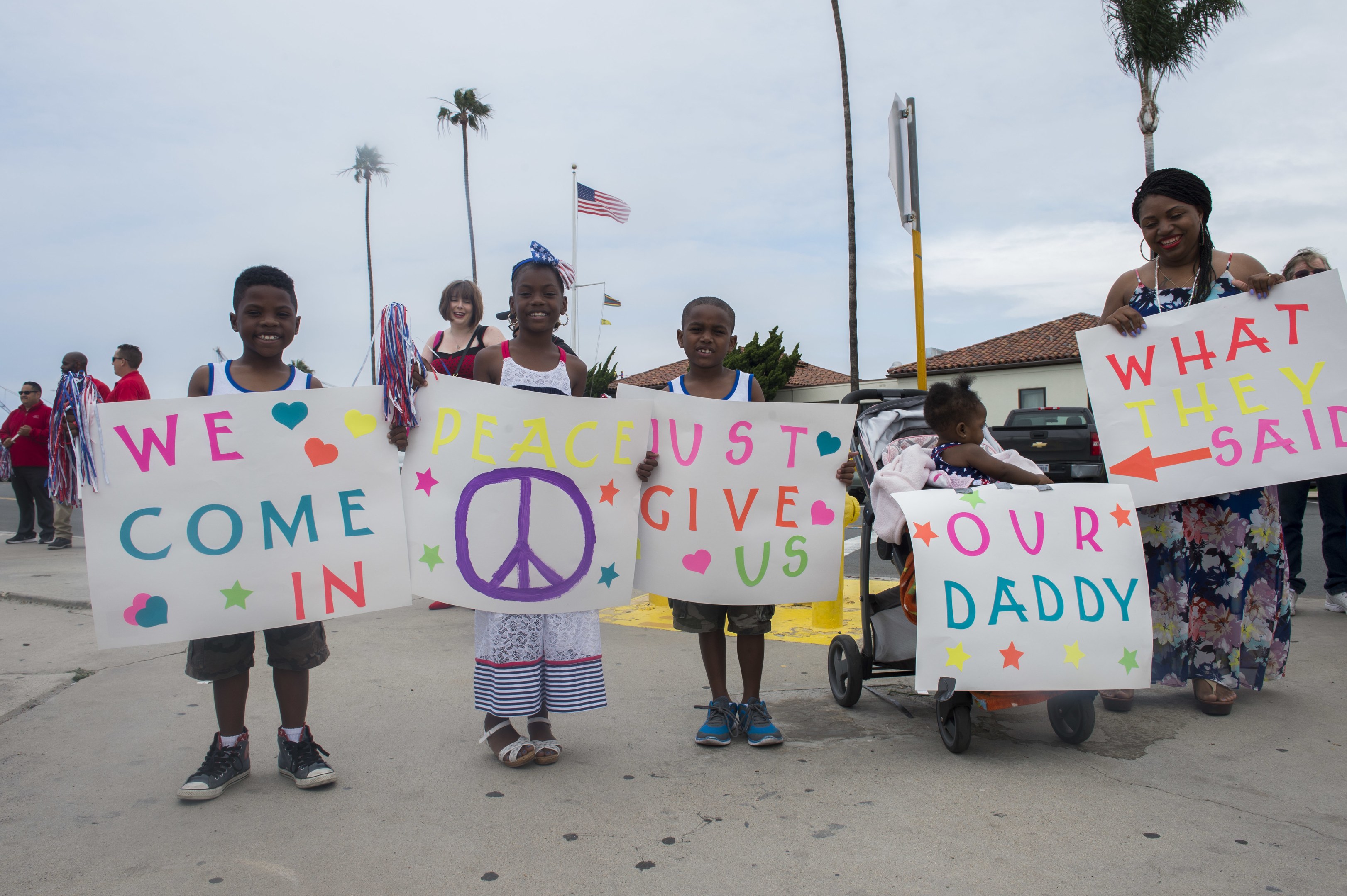 Eine Gruppe von Kindern hält Schilder mit der Aufschrift "Wir kommen in Frieden, gebt uns unseren Daddy" vor einem Gebäude, mit einer Frau, die einen Kinderwagen schiebt, umgeben von Bäumen, Pfählen, einer Flagge, einem Haus, Pflanzen und einem bewölkten Himmel.