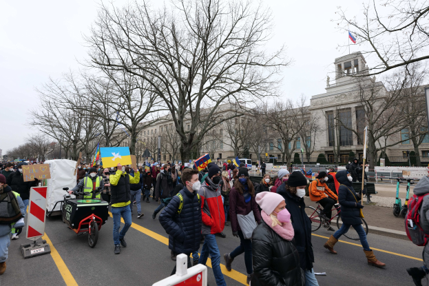Eine große Gruppe von Menschen marschiert auf einer Stadtstraße, einige halten Schilder und andere fahren Fahrräder, mit Bäumen und einem Gebäude im Hintergrund unter einem klaren blauen Himmel.