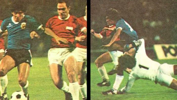 Eine Gruppe von Männern, die auf einem Feld Fußball spielen, mit einem Scoreboard im Hintergrund, ein Spieler tritt den Ball, während andere versuchen, die Kontrolle zu erlangen.