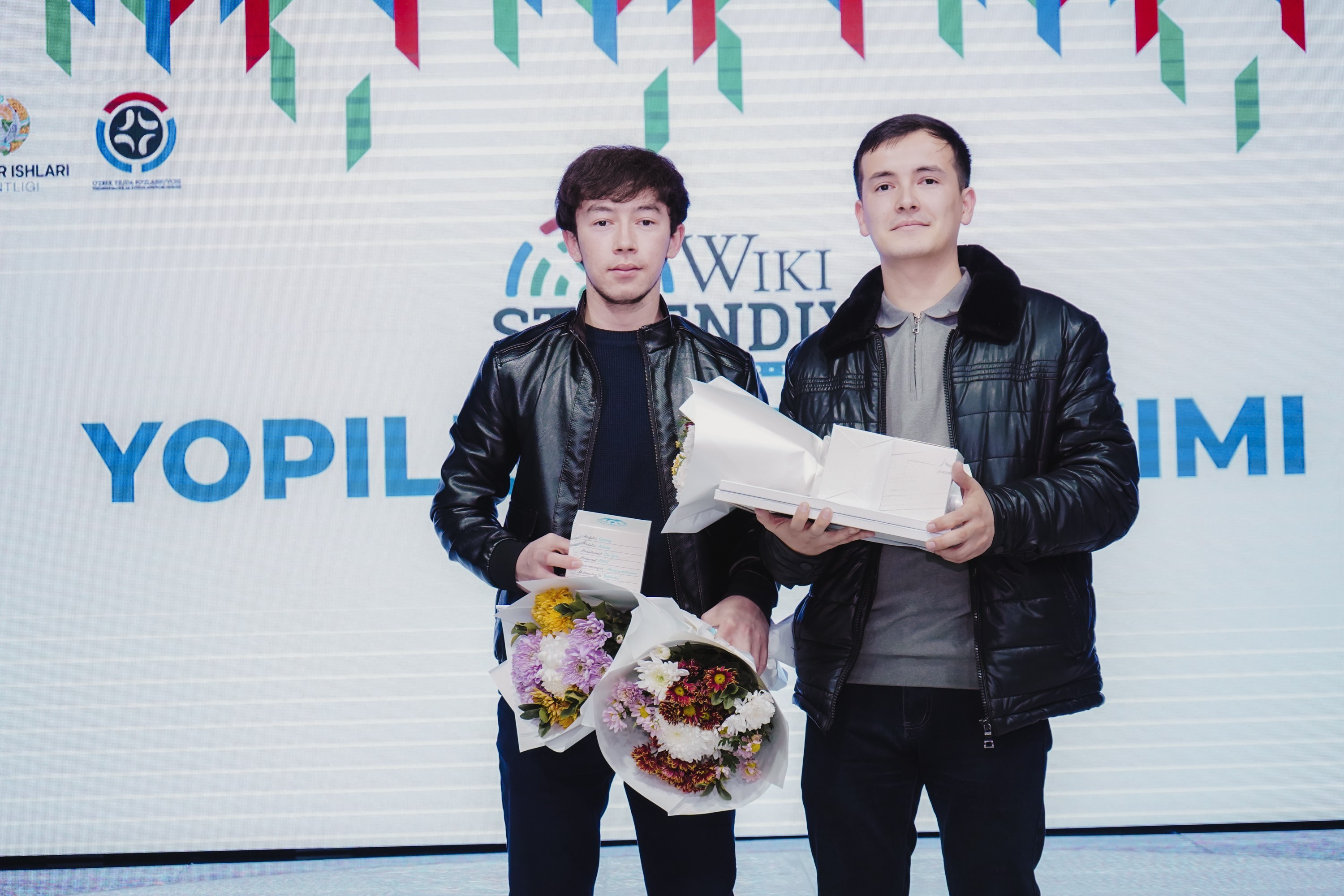 Zwei Männer in schwarzen Jacken stehen auf einer Bühne und halten Blumen, mit einem 'Yopil International Film Festival'-Banner im Hintergrund.
