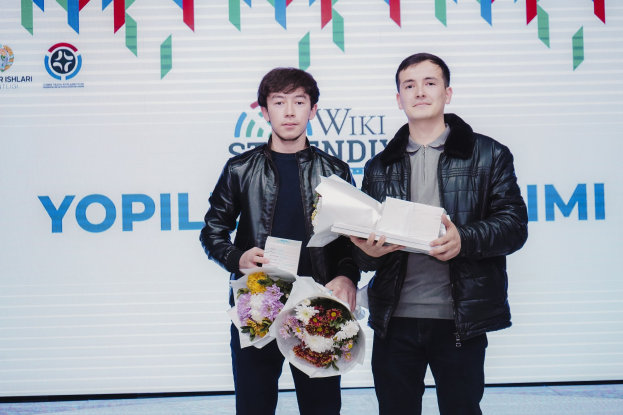 Zwei Männer in schwarzen Jacken stehen auf einer Bühne und halten Blumen, mit einem 'Yopil International Film Festival'-Banner im Hintergrund.