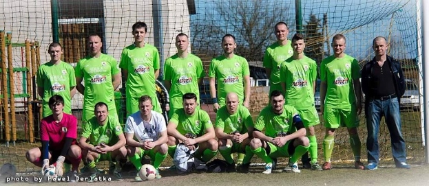 Eine Gruppe von Männern in grünen T-Shirts steht auf einem Fußballfeld mit einem Ball vor ihnen, einige in der Hocke, in der Nähe eines Torpfostens, mit Bäumen, Gebäuden und Fahrzeugen im Hintergrund.