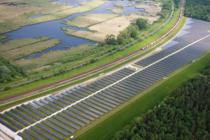 Luftaufnahme einer großen Solar-Farm mit Solar-Panelen, umgeben von Bäumen, Gras, Pflanzen und Wasser, mit einem Zug auf einer nahen Bahnstrecke.