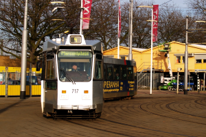 Weiß-blauer Tram auf einer Stadtstraße mit einem Passagier im Inneren, umgeben von Laternen, Bannern, einem Zaun, anderen Fahrzeugen, Gebäuden, Bäumen und einem klaren blauen Himmel.