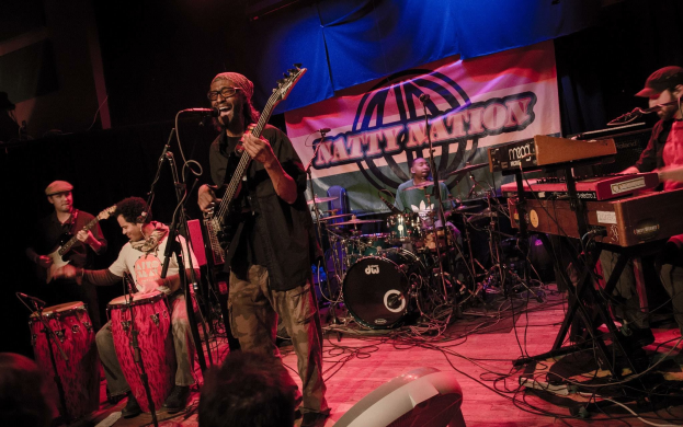 Eine Live-Band performt auf der Bühne mit Mikrofonen und Ständern, begleitet von einem Publikum, unter einem Banner in einer abgedunkelten Umgebung.