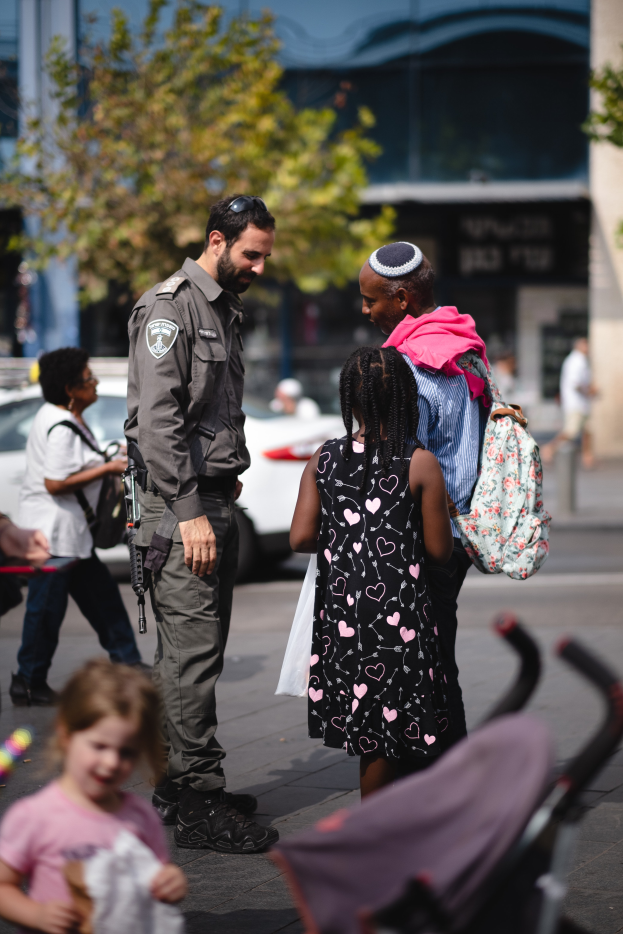 Ein Polizist spricht zu einer Gruppe von Menschen auf der Straße, mit einem Kinderwagen im Vordergrund und Fahrzeugen, Bäumen und einem Gebäude im Hintergrund.
