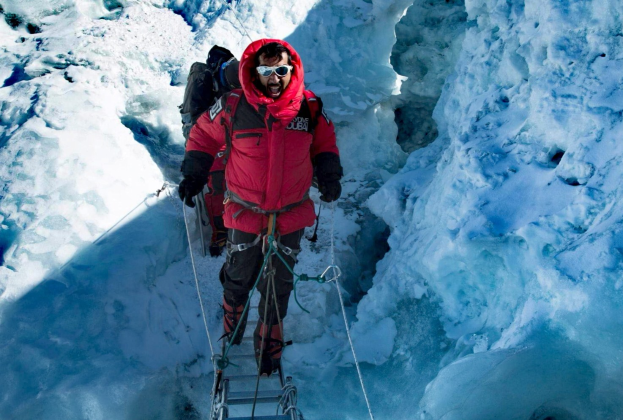 Ein Mann in einer roten Jacke klettert einen schneebedeckten Berg hoch, ausgestattet mit einer Brille, Handschuhen, einem Rucksack und Seilen.