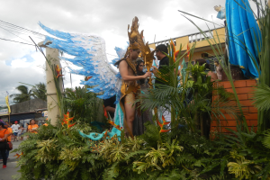 Eine Gruppe von Menschen geht neben einem Karnevalswagen mit Pflanzen und Blumen Verzierung, mit kostümierten Personen auf dem Wagen, Gebäuden, Bäumen und Strommasten mit Drähten im Hintergrund unter einem bewölkten Himmel.