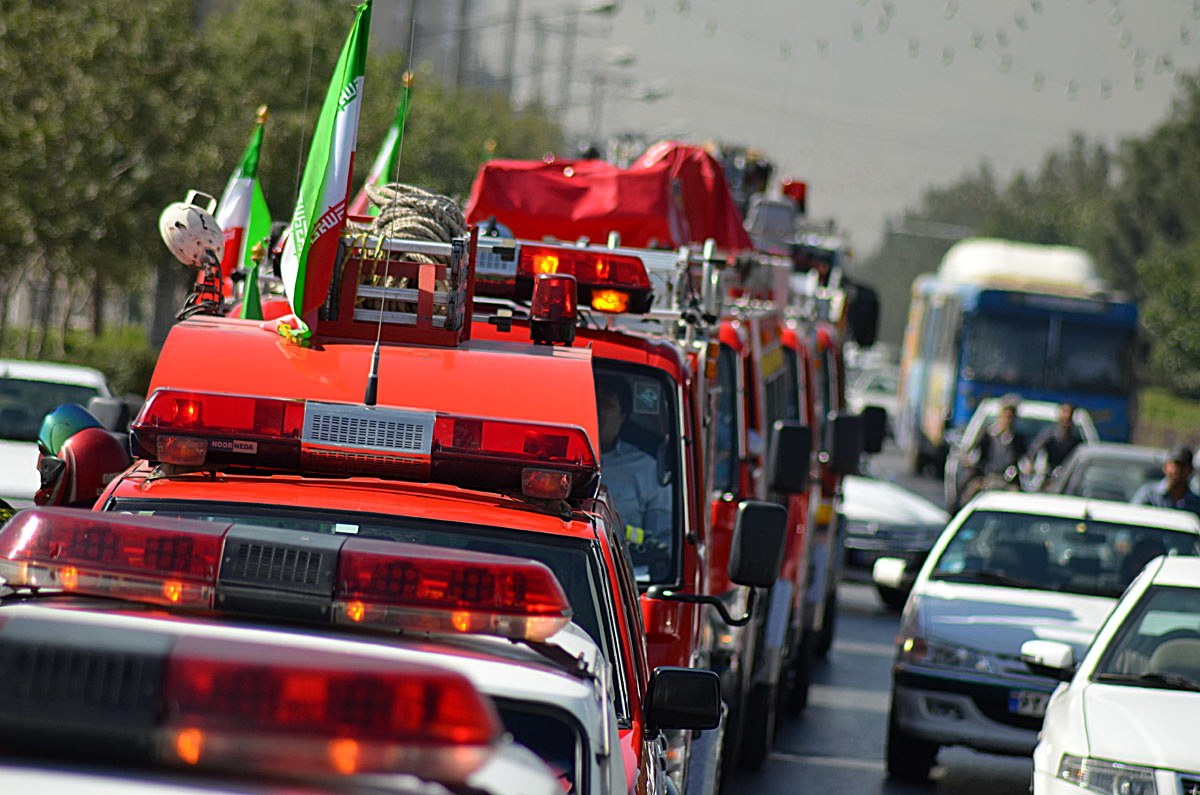 Busy street with cars, trucks, and a fire truck with flags, people in vehicles, trees, poles, and a clear blue sky in the background.
