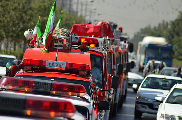 Busy street with cars, trucks, and a fire truck with flags, people in vehicles, trees, poles, and a clear blue sky in the background.