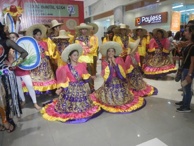 Eine Gruppe von Menschen in bunten Kleidern und Hüten, die in einem Einkaufszentrum während eines festlichen Ereignisses tanzen, während andere Musikinstrumente spielen und ein Banner im Hintergrund zu sehen ist.