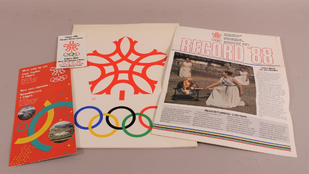 Eine weiße Fläche zeigt eine Sammlung von Büchern und Broschüren mit den Olympischen Ringen, Texten und Bildern zum Thema Olympische Winterspiele 1988.