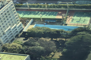 Eine Luftaufnahme eines Tennisplatzes in einer Stadt, umgeben von Gebäuden, Bäumen, Pflanzen, Pfählen, Fahrzeugen und einem Schwimmbad.