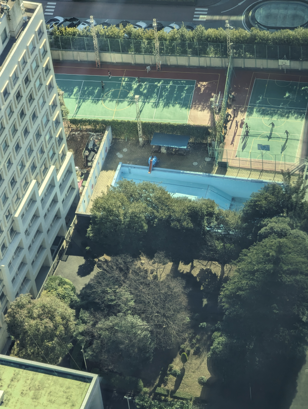 Eine Luftaufnahme eines Tennisplatzes in einer Stadt, umgeben von Gebäuden, Bäumen, Pflanzen, Pfählen, Fahrzeugen und einem Schwimmbad.