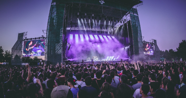 Eine große Menschenmenge auf einem Musikfestival schaut Künstlern auf einer beleuchteten Bühne mit Instrumenten und Bildschirmen zu, die vor einem Gebäude, Bäumen und einem klaren blauen Himmel aufgebaut ist.