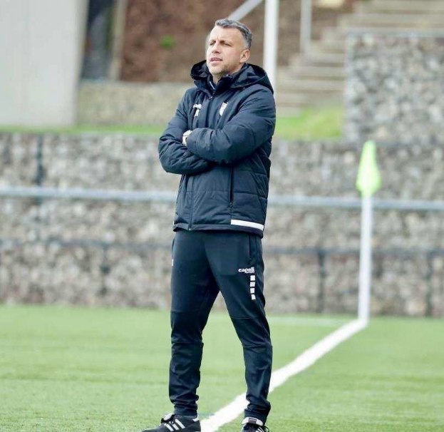 Ein Mann in schwarzer Jacke und Hose steht auf einem Fußballfeld mit verschränkten Armen, sieht aus wie ein Trainer während eines Trainings.