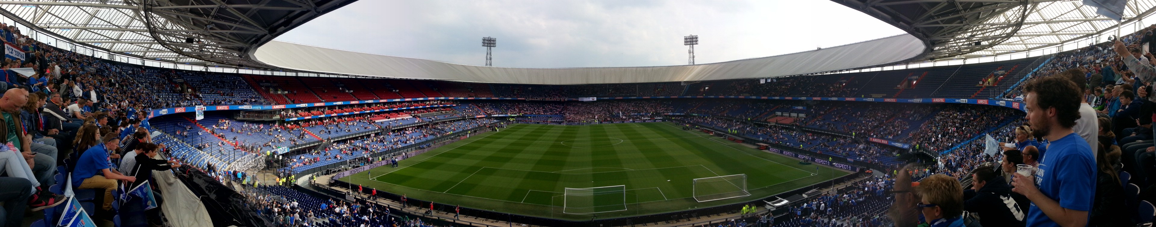 Ein volles Stadion mit Zuschauern bei einem Fußballspiel, mit Menschen in den Rängen, die Kameras halten, ein Netz auf dem Boden, Lichtmasten im Hintergrund und einen klaren blauen Himmel.