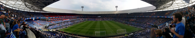 Ein volles Stadion mit Zuschauern bei einem Fußballspiel, mit Menschen in den Rängen, die Kameras halten, ein Netz auf dem Boden, Lichtmasten im Hintergrund und einen klaren blauen Himmel.