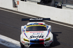 Ein BMW M6 GT3 Rennauto auf einer Rennstrecke mit Text auf der Karosserie, eine Wand im Hintergrund und Maschendrahtzaun.