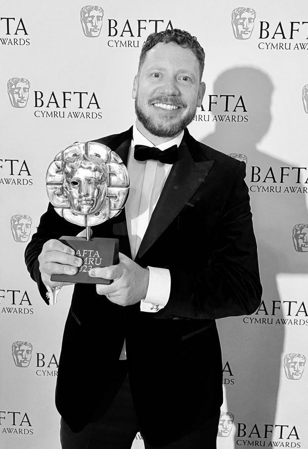Ein Mann in einem Smoking, der eine BAFTA-Trophäe hält und lächelt, mit einem Hintergrundplakat, auf dem "BAFTA Awards 2018" steht