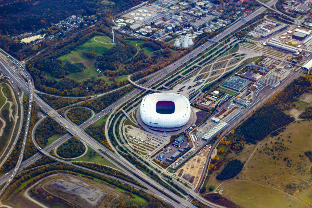 Luftaufnahme des Olympiastadions in München, Deutschland, mit umliegenden Gebäuden, Bäumen, Straßen, Fahrzeugen und Gras.