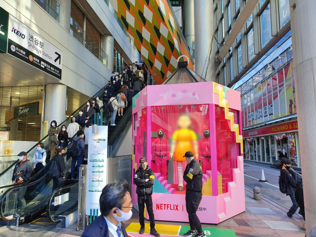 Eine Gruppe von Menschen steht vor einem gläsernen Gebäude mit Texttafeln, einige tragen Masken, während eine Rolltreppe mit Menschen daneben fährt; in der Mitte eine pinkfarbene Box mit der Aufschrift 'Netflix's new Netflix Booth in Tokyo'.