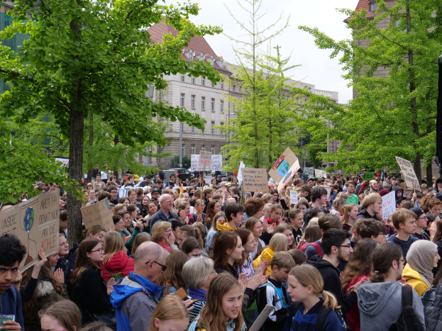 Eine große Menge von Menschen mit Schildern steht vor einem Gebäude in Berlin, mit Bäumen, Fahrzeugen und einem Lautsprecher, unter einem klaren Himmel.