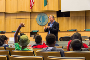 Ein Mann in einem schwarzen Blazer, blauem Hemd und schwarzen Hosen steht vor einer Klasse und h├Ąlt ein Mikrofon in der Hand, mit Kindern, die aufmerksam zuh├Âren, einer h├Âlzernen Wand mit Logo und zwei Flaggen dahinter und einem wei├čen Bildschirm rechts.