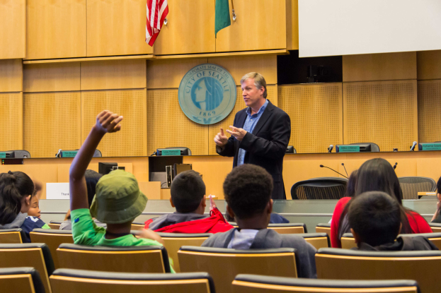Ein Mann in einem schwarzen Blazer, blauem Hemd und schwarzen Hosen steht vor einer Klasse und h├Ąlt ein Mikrofon in der Hand, mit Kindern, die aufmerksam zuh├Âren, einer h├Âlzernen Wand mit Logo und zwei Flaggen dahinter und einem wei├čen Bildschirm rechts.