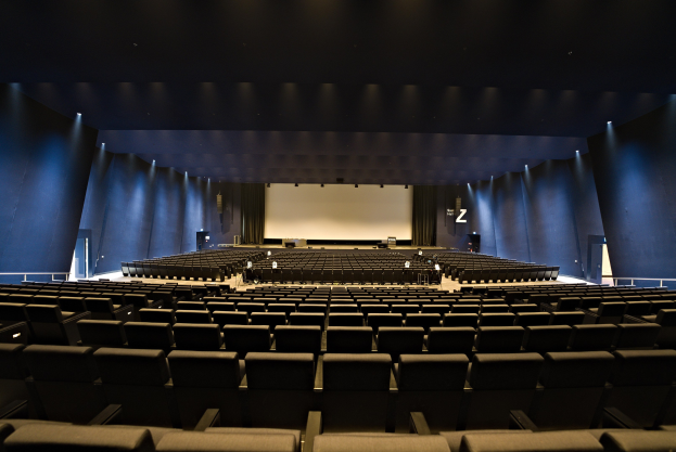 Ein großer Zuschauerraum mit Reihen von Stühlen, wandmontierten Lampen, einem Projektionsschirm und ein paar Gegenständen auf der Bühne.