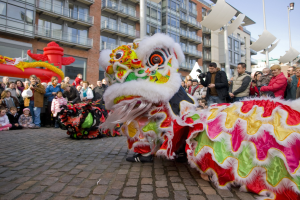Ein lebendiges chinesisches Neujahrsfest in Amsterdam mit einem Löwen tanzen im Vordergrund mit einer Menschenmenge, einige halten Kameras, und Gebäude, Laternenmasten und einen klaren blauen Himmel im Hintergrund.