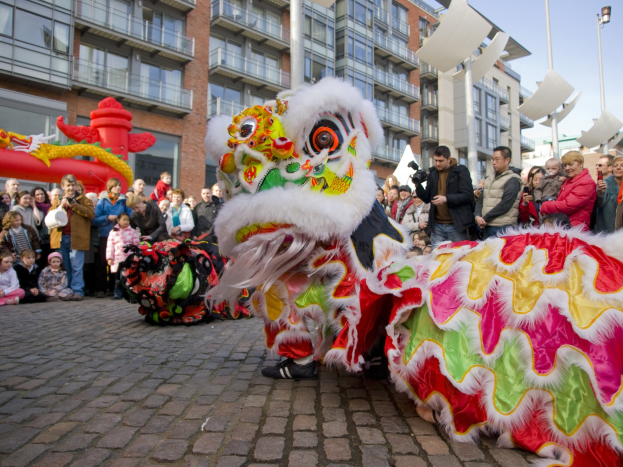 Ein lebendiges chinesisches Neujahrsfest in Amsterdam mit einem Löwen tanzen im Vordergrund mit einer Menschenmenge, einige halten Kameras, und Gebäude, Laternenmasten und einen klaren blauen Himmel im Hintergrund.