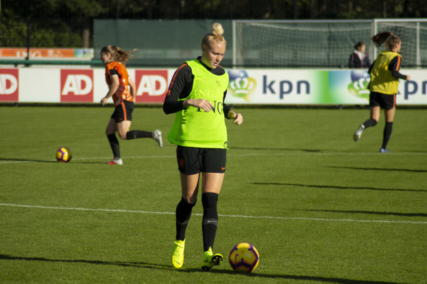 Gruppe von Frauen, die Fußball auf einem Feld mit Bäumen im Hintergrund, Bannern mit Text und einem Netz im Vordergrund spielen.