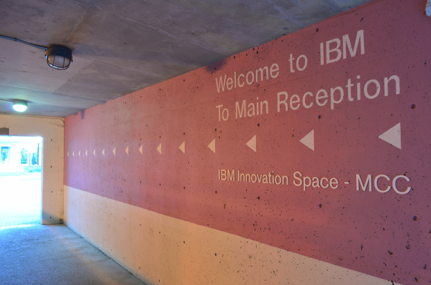 Eine Wand mit dem Text "Willkommen bei IBM" beleuchtet von Deckenlampen, mit einer Tür im Hintergrund beschriftet mit "IBM Innovationsraum MCC."