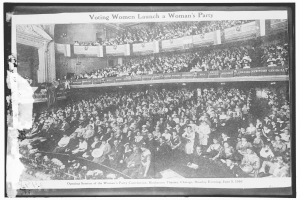 Ein Schwarz-Wei├č-Foto einer gro├čen Menge von Menschen in einem Geb├Ąude, einige sitzen, einige stehen, Schilder mit Text und eine Wand im Hintergrund. Unten steht der Text "Wahlberechtigte Frauen gr├╝nden eine Frauenpartei."